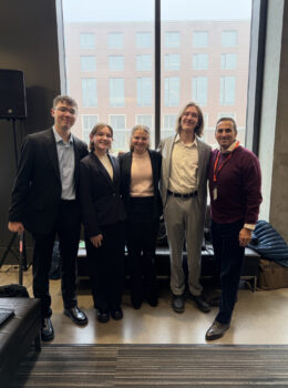Five-member sales team in Oregon standing indoors in front of a large window with a brick office building visible outside; four young professionals in business attire and one older man in a sweater, all smiling and posing for a group photo.