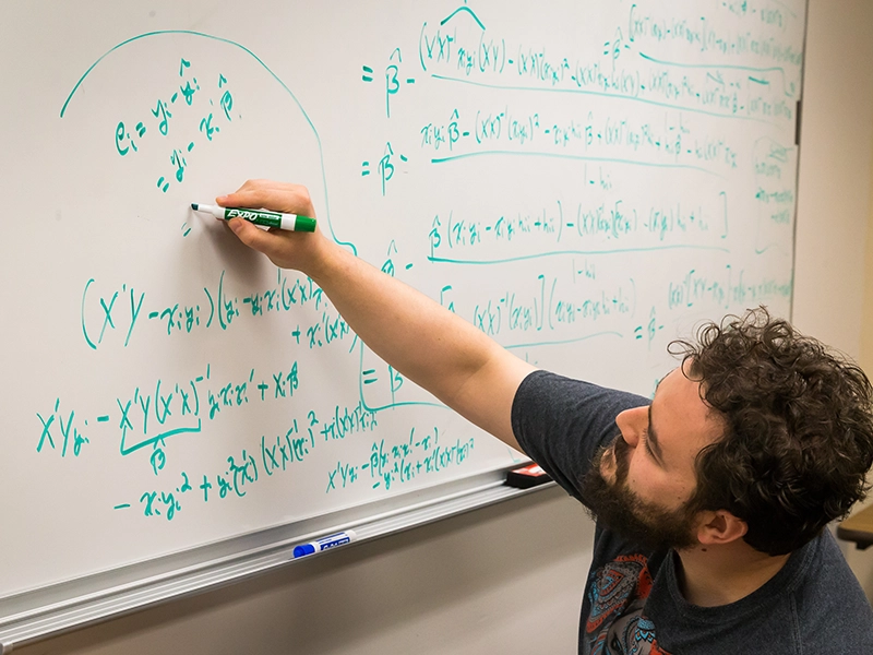 Graduate student solving advanced statistical equations on a whiteboard with a green marker.