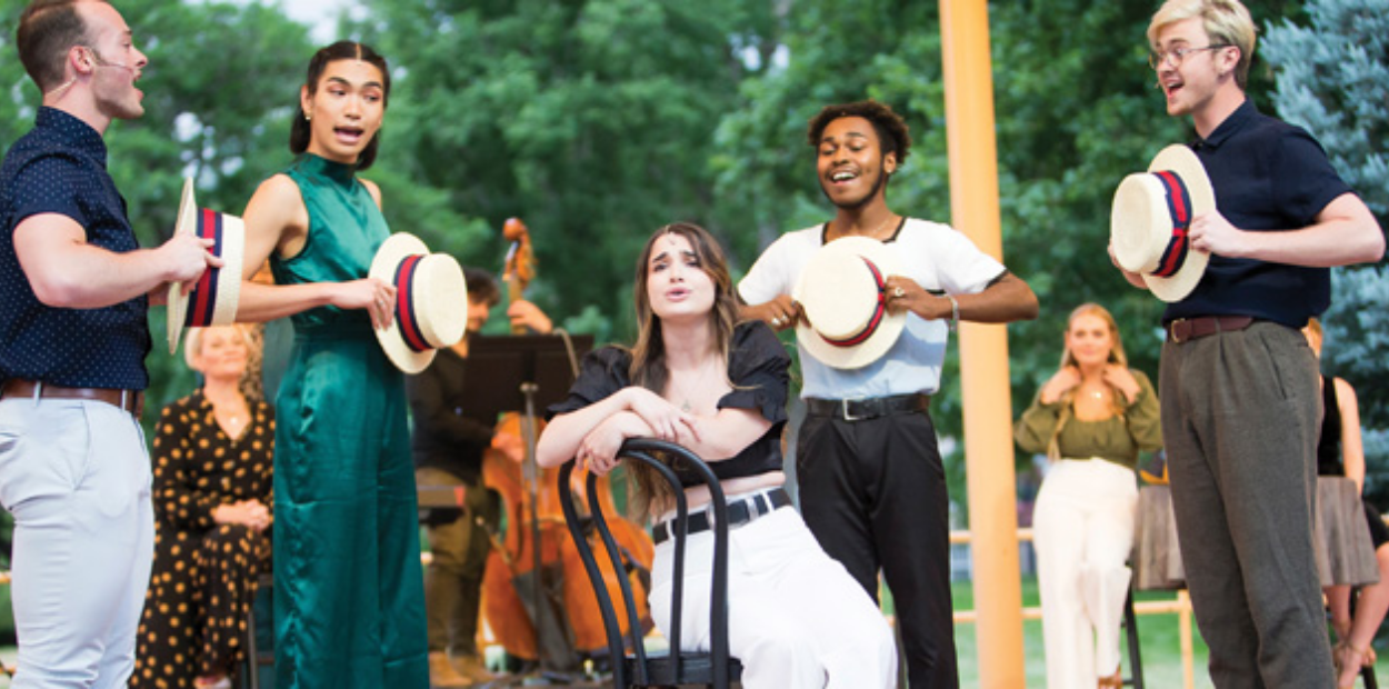 A group of performers standing on an outdoor stage, singing and holding barbershop quartet hats.