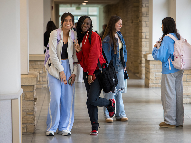Two students posing for a photo while two more students have a conversation in the background.
