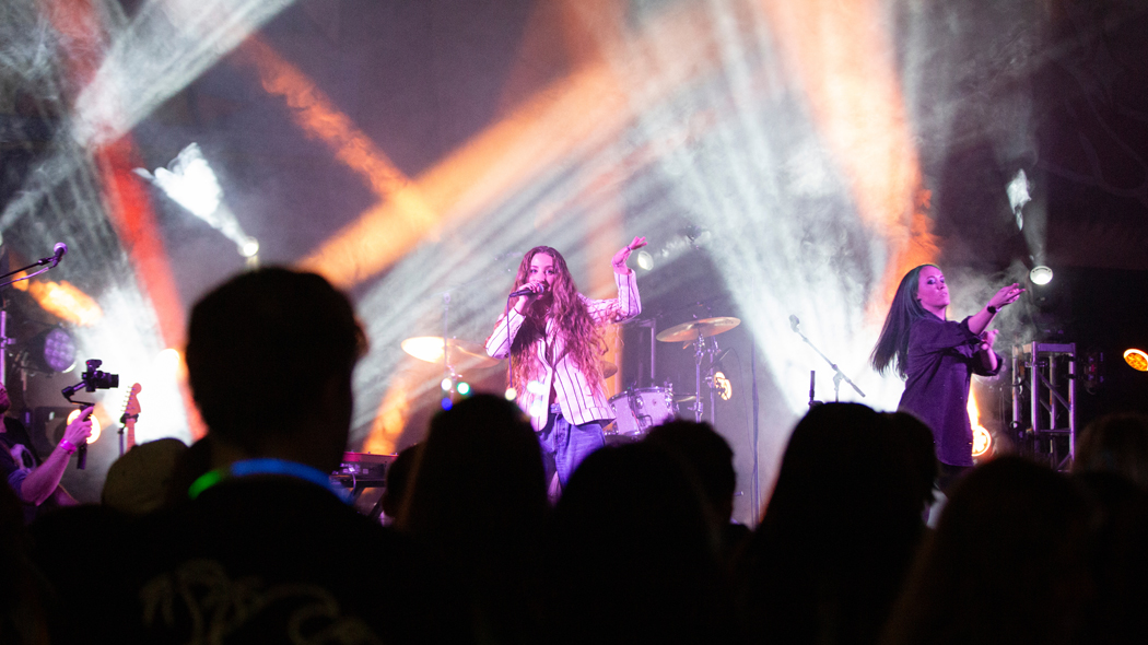 Singer performing on stage with colorful lights in the background and crowd in silhouette in the foreground