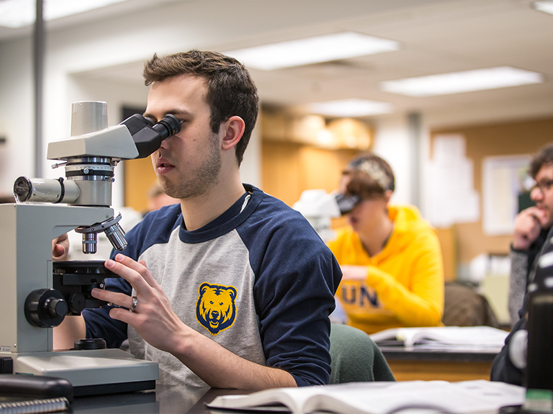 Student in classroom looking at slides through a microscope with other students in the background.