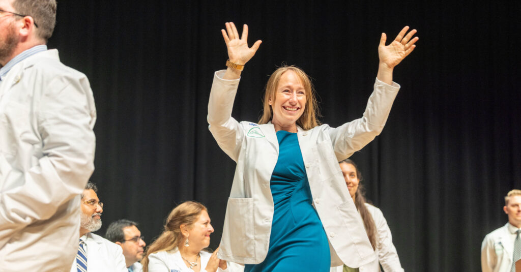 A medical student smiling with her hands in the air wearing her white coat