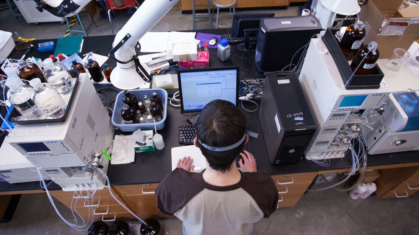 Chemistry student in lab looking at computer screen while surrounded by equipment.