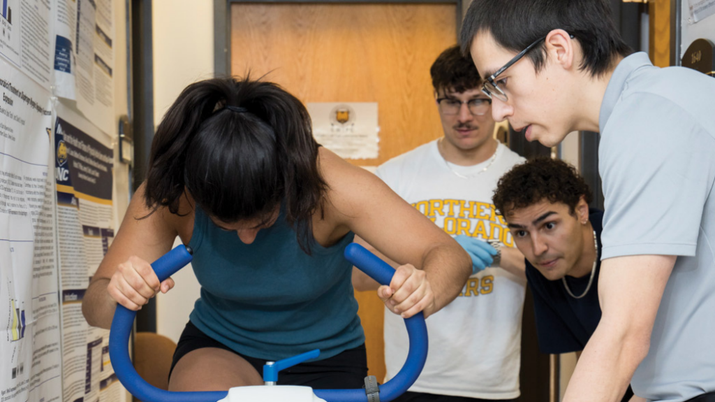 A woman riding a stationary bike while three onlookers observe to collect data.