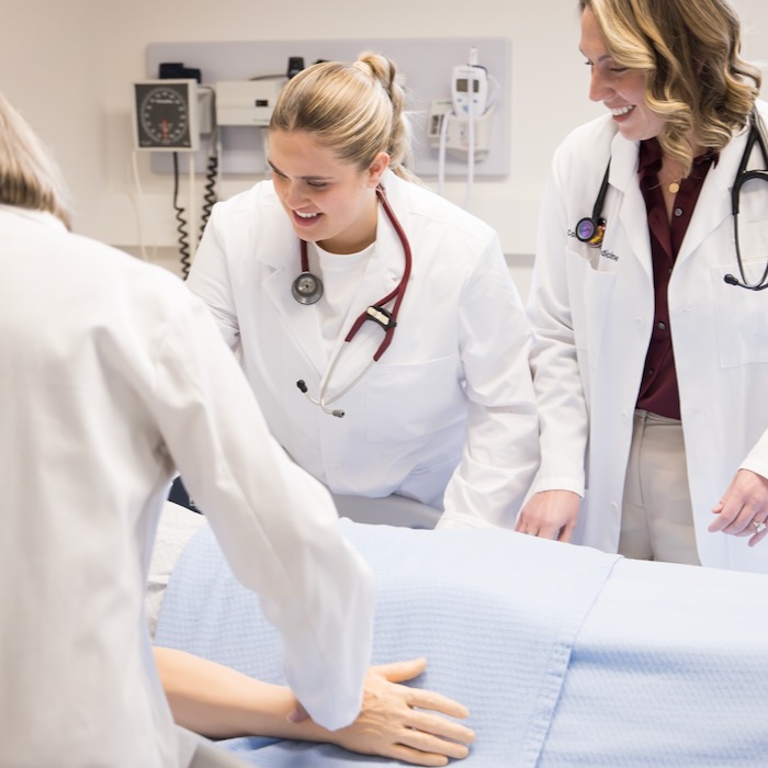 A student and two supervising physicians are smiling while talking to a patient in bed.