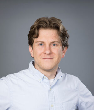 Headshot of Kyle Oswald. A person with light brown hair and a light blue button-up shirt smiles slightly against a plain gray background.