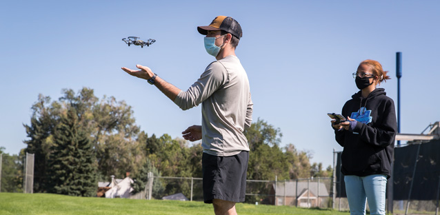 A person having a drone hovering over his hand wearing a hat and a mask outside