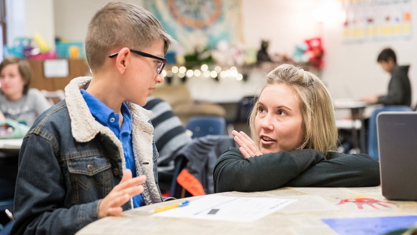 Teacher talking to a young student at a table in a classroom.