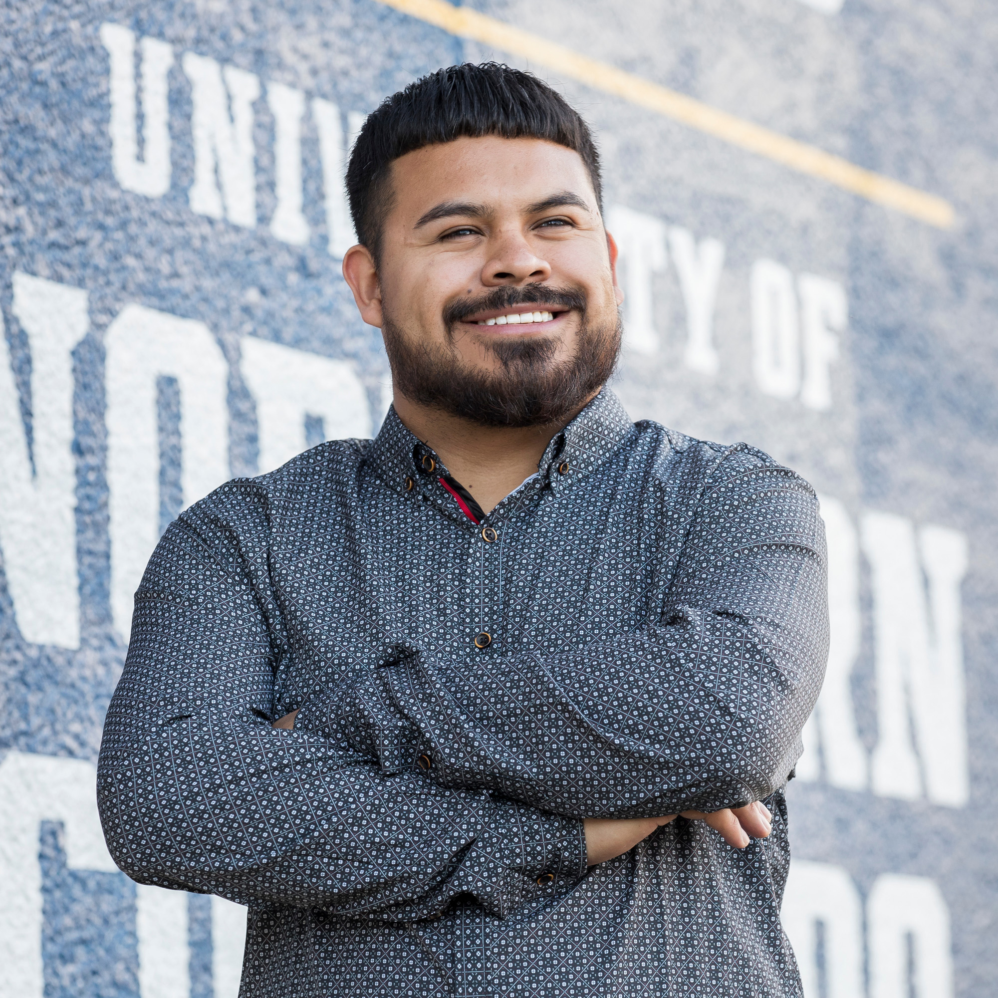 UNC graduate, Fernando Beltran smiling with his arms crossed confidently in front of a painted UNC logo.