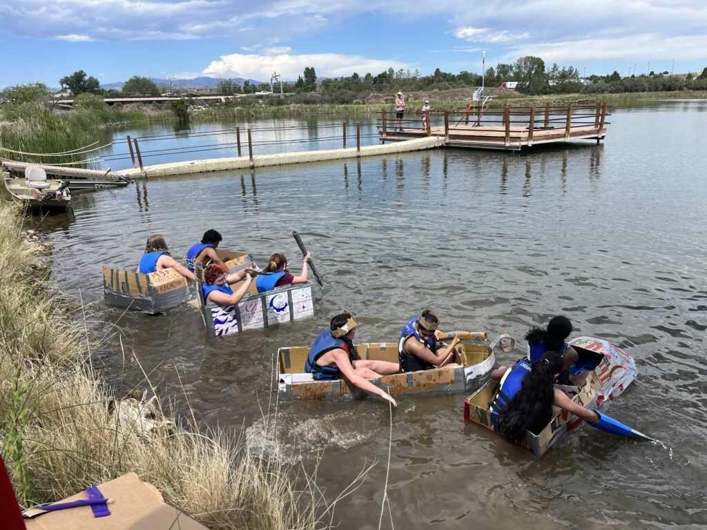 Student participants of the Frontiers of Science Institute paddling cardboard boats across a lake.