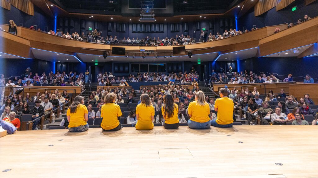 Six UNC students wearing yellow shirts with their back toward the camera, sitting on a stage facing many students sitting in chairs