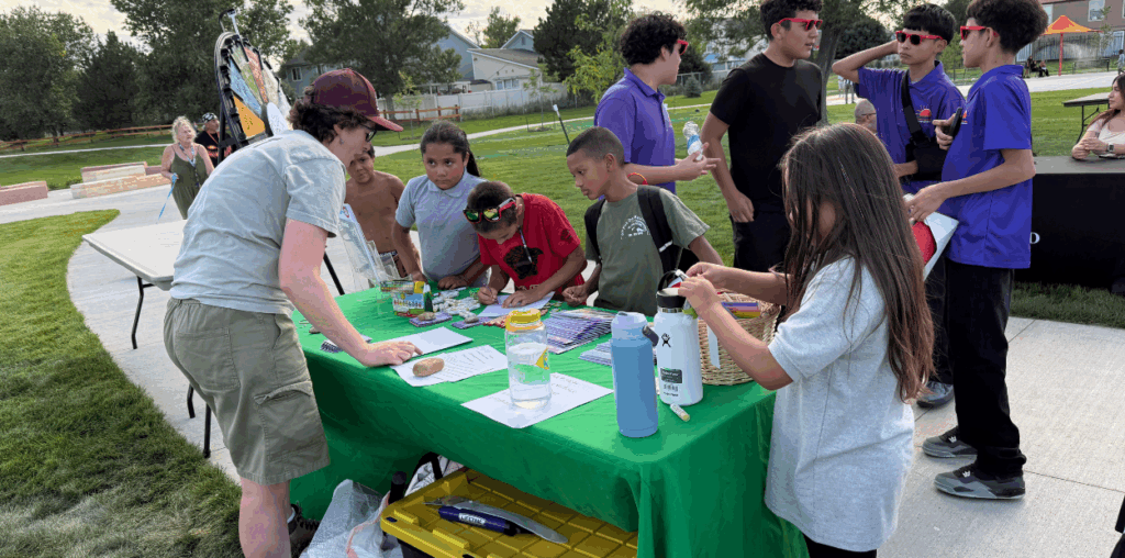 A group of students standing near a green table talking to an adult standing on the opposite side