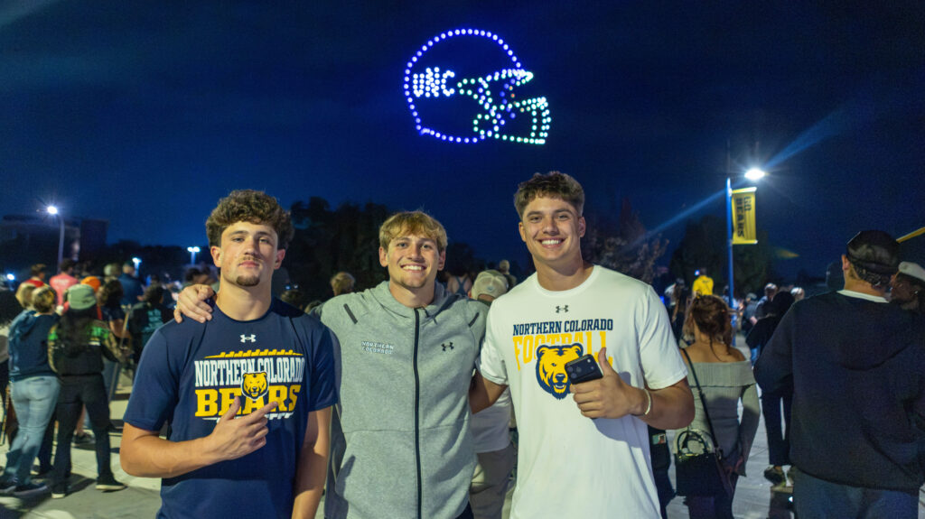 Three UNC students smiling in front of a UNC football helmet lit up by drones in the sky