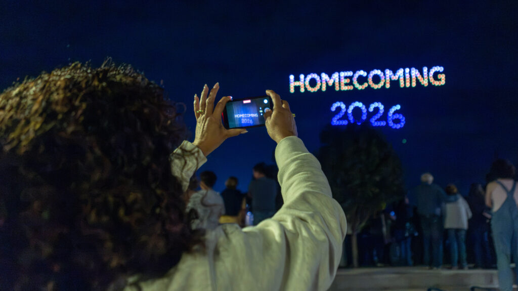 A UNC student taking a picture of the words Homecoming 2026 lit up in the sky by drones