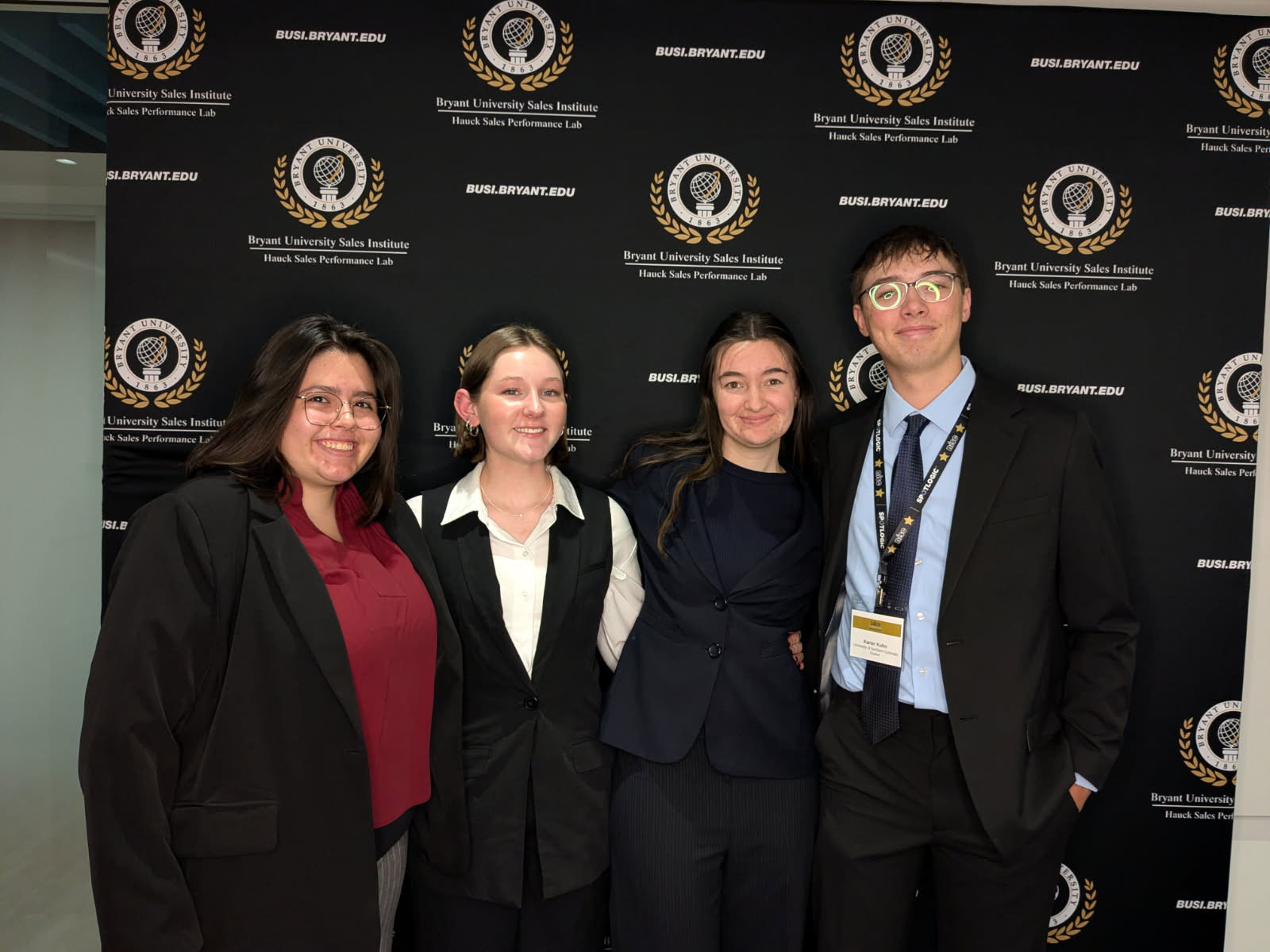 Four people are posing together, smiling, standing in front of a backdrop displaying the Bryant University Sales Institute logo. They are dressed in professional attire.