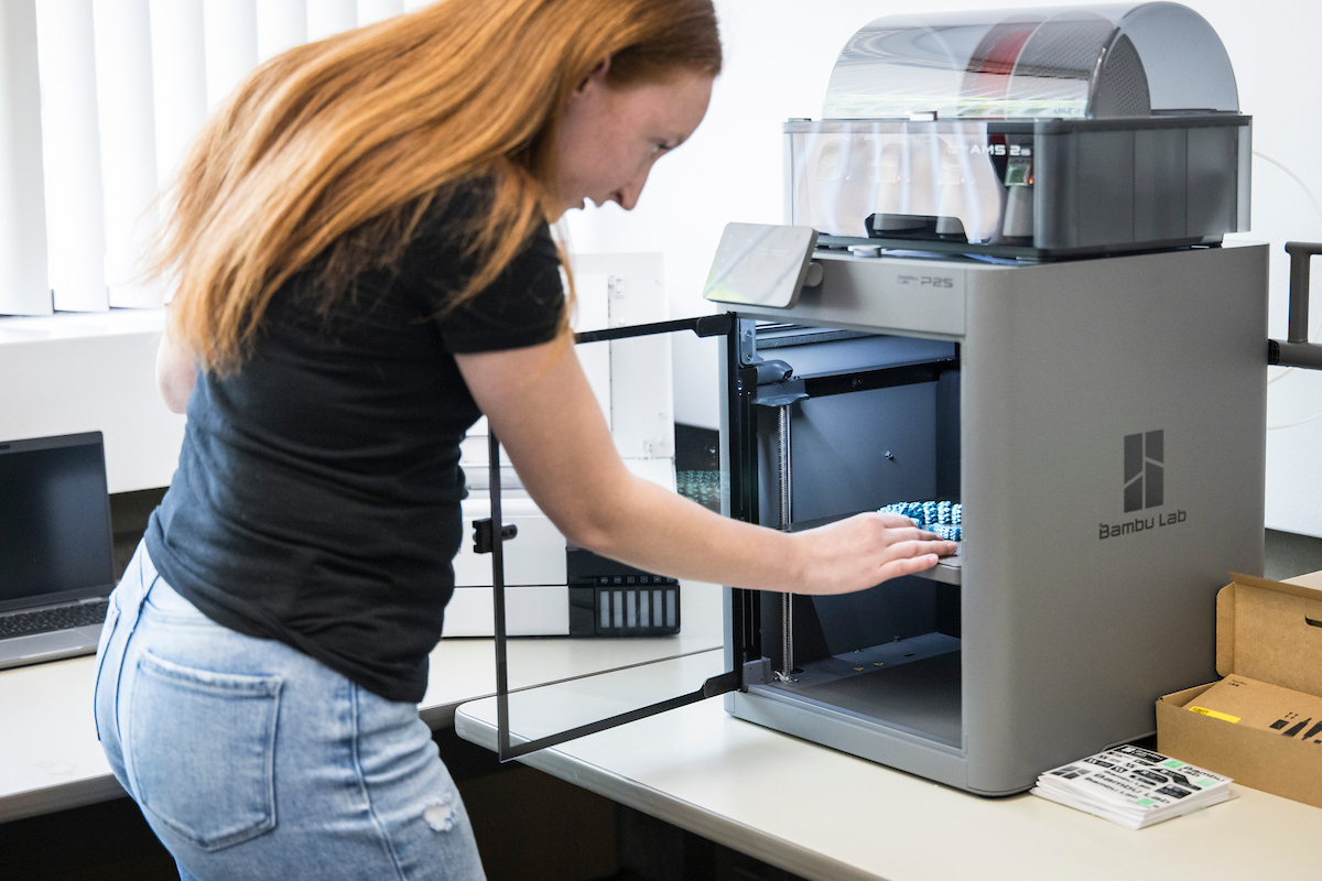 A student stands in front of a 3D printer with the door open, looking at an object inside