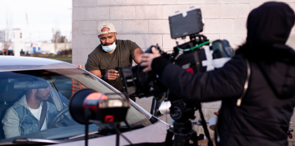 Three UNC students working on a film set, one in a car, one filming and the third giving instructions as the director.