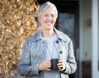 Kathy Zellers standing in front of a tree smiling, and holding a black coffee mug