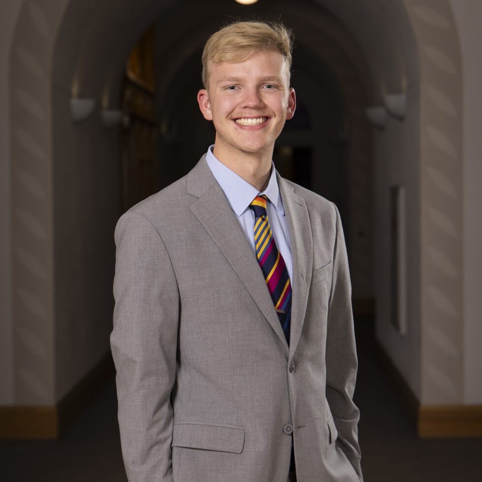 A headshot of Keller McBride wearing a brown suit