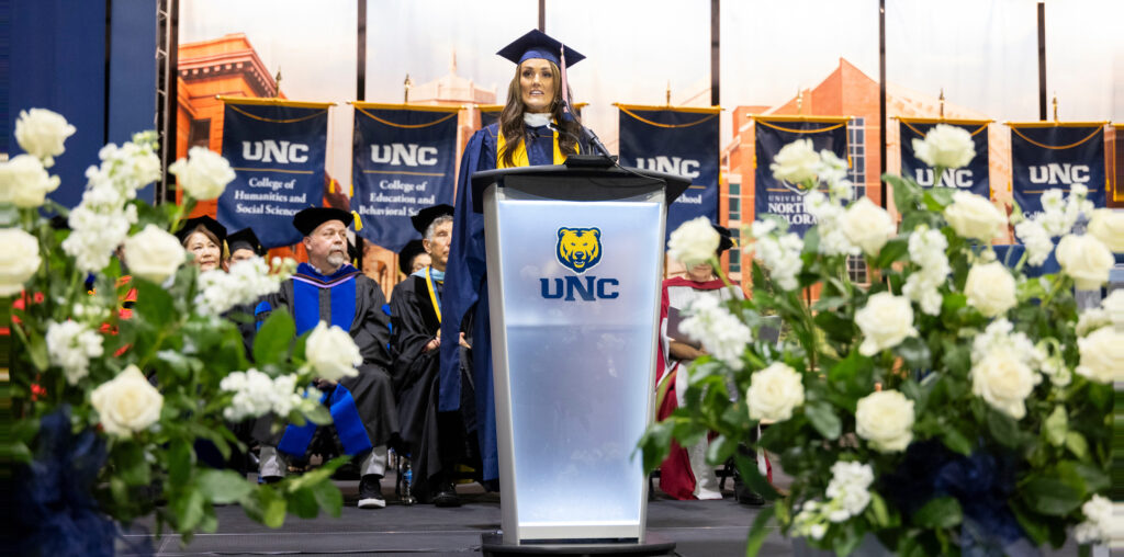 Kim Corban standing at a podium in her commencement gown giving a speech on stage with white flowers surrounding the stage