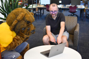 UNC mascot Klawz, wearing a yellow shirt, sits at a table with a librarian. The librarian is pointing to information on a laptop screen.