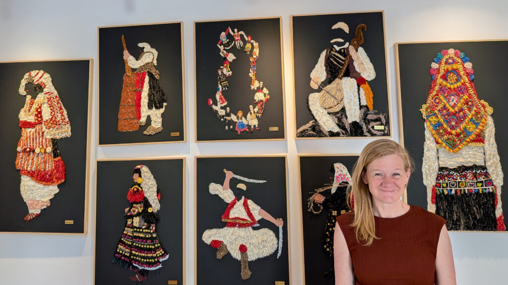 Britney Kyle in front of an art display showing traditional Albanian costumes made from pieces of plants