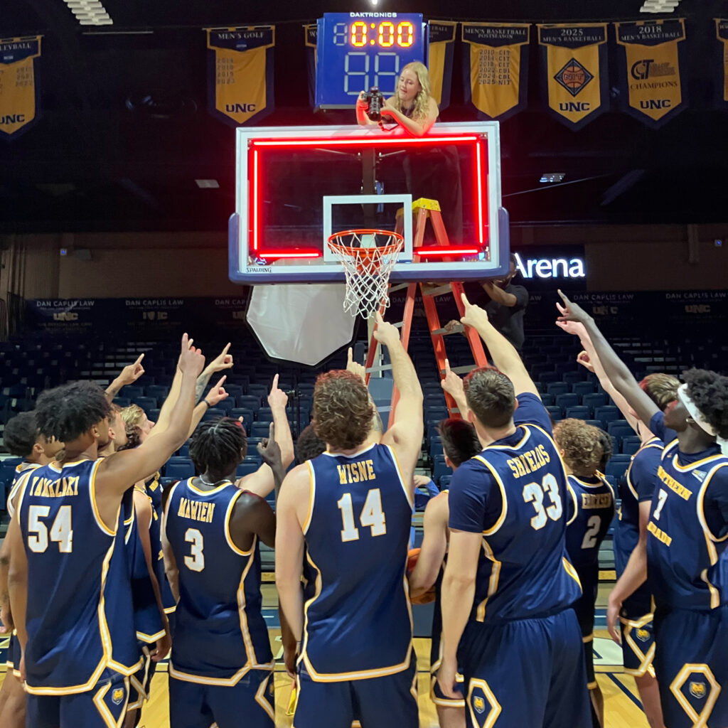 Lexie Martin taking a picture from the top of a basketball hoop with the UNC men's basketball team pointing toward her on the ground