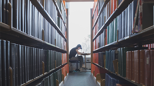 Bookshelves in Michener library with student studying at table in the background.