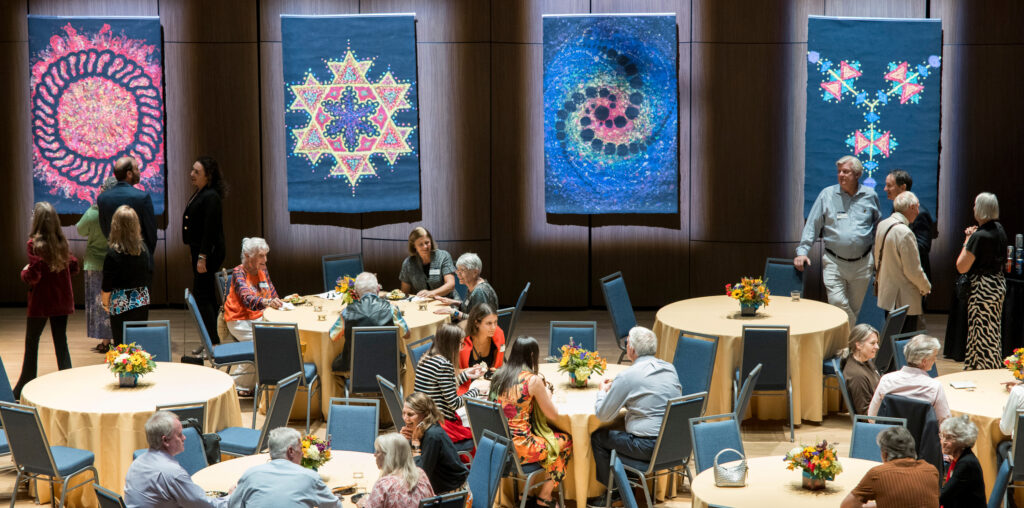 A group of people sitting at tables with Lydia Ruyle's art on the wall