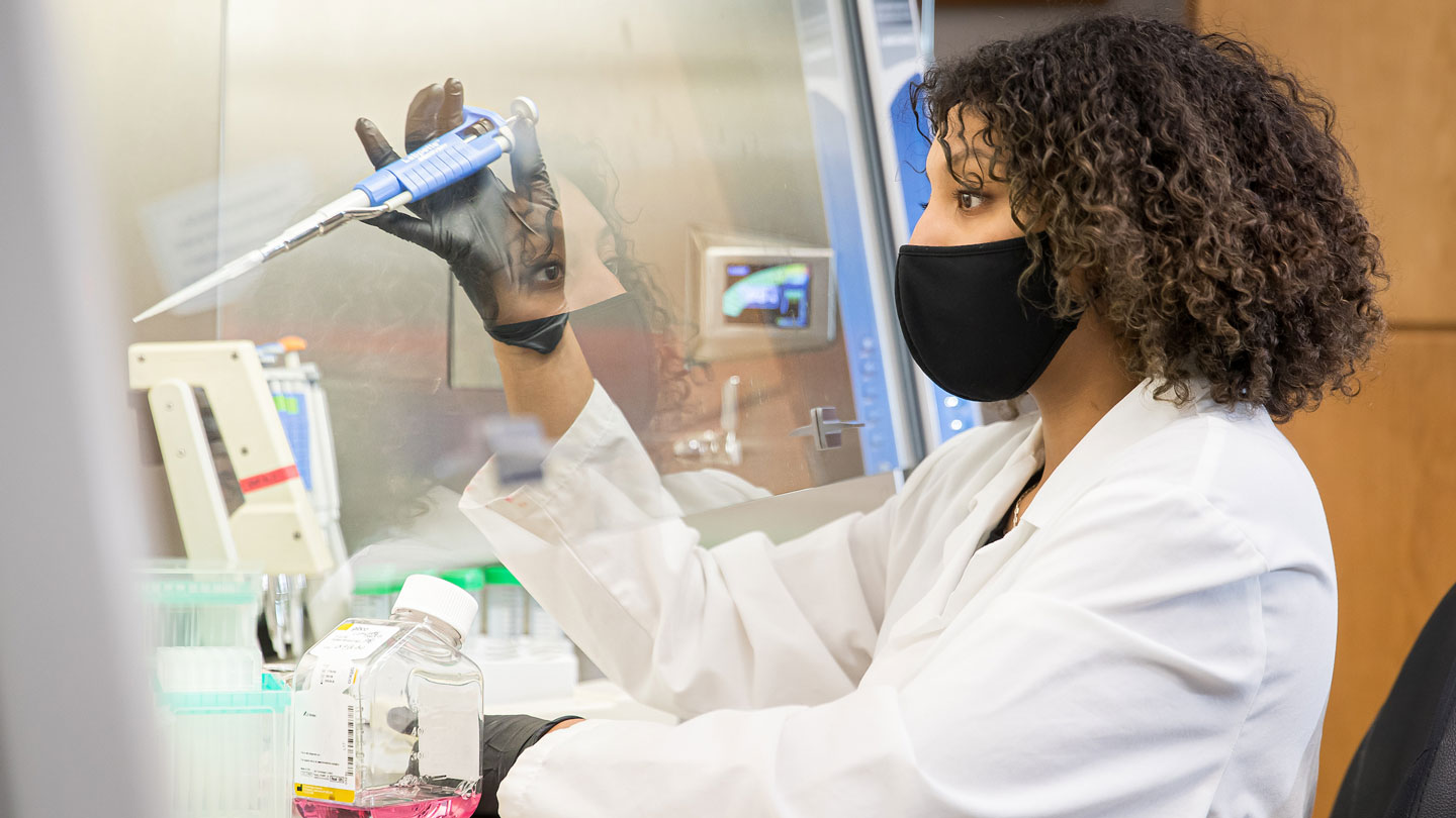Student in mask and lab coat behind glass, using a scientific dropper.