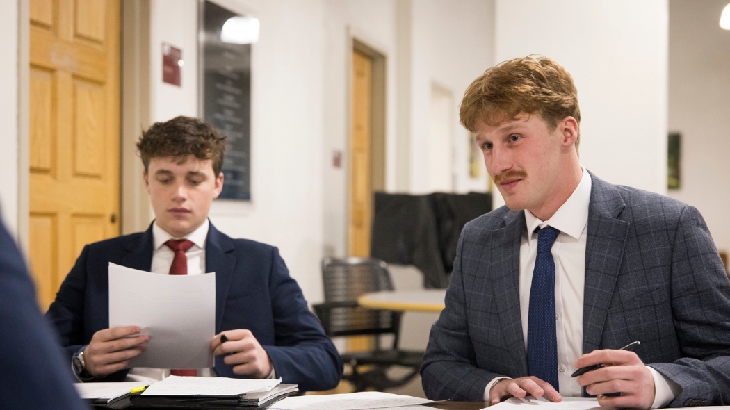 Two students in suits talking to a competition judge across a table.