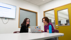 Two people seated at a table in a Michener Library study room look at a laptop on a table. There is a water bottle next to the laptop.