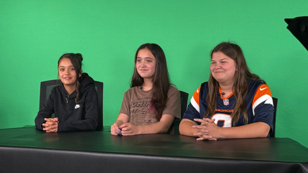 Three University Middle School students sitting at a desk in front of a green screen