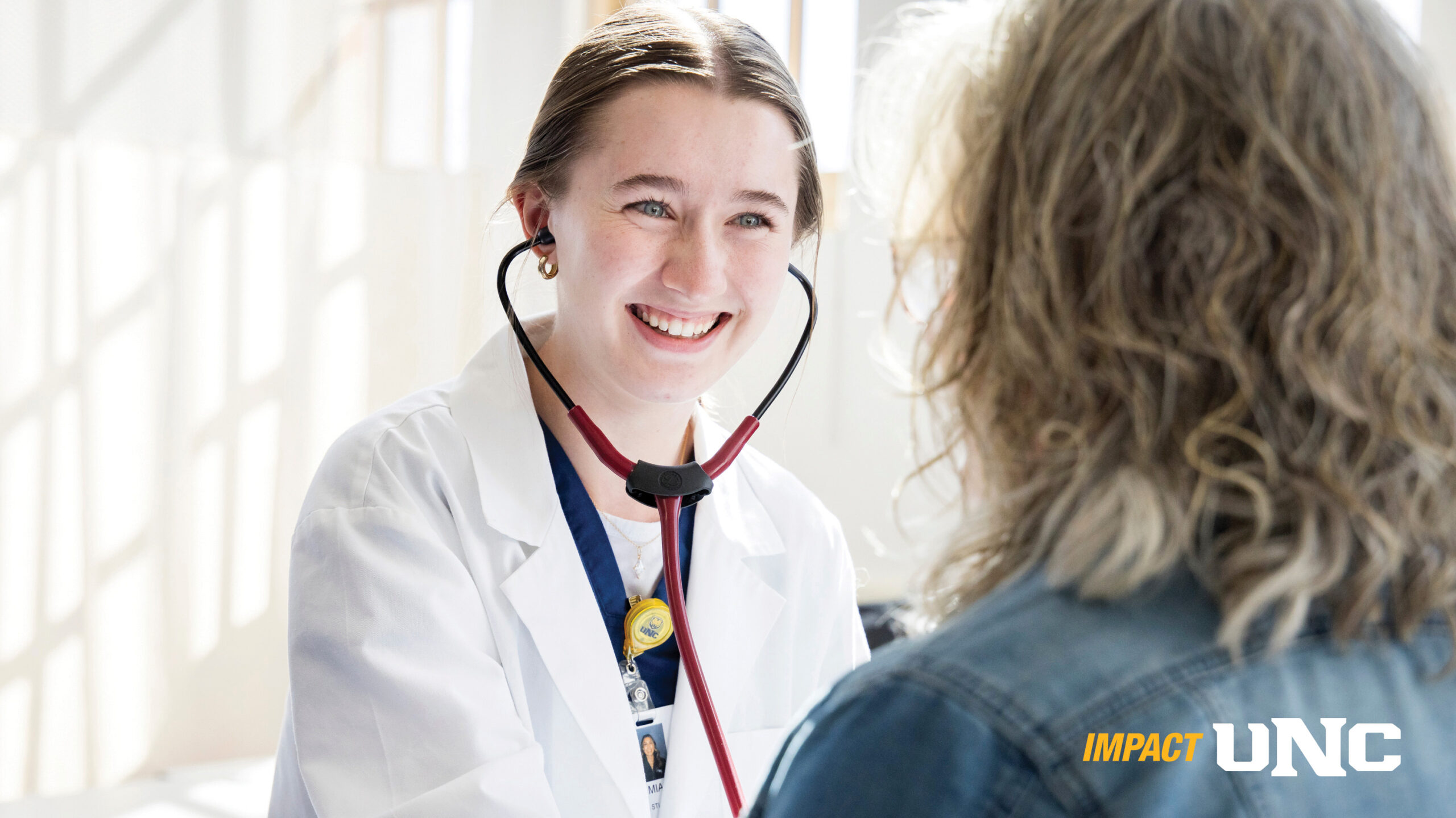 A UNC nursing student wearing a white jacket and a stethoscope, smiling at a patient