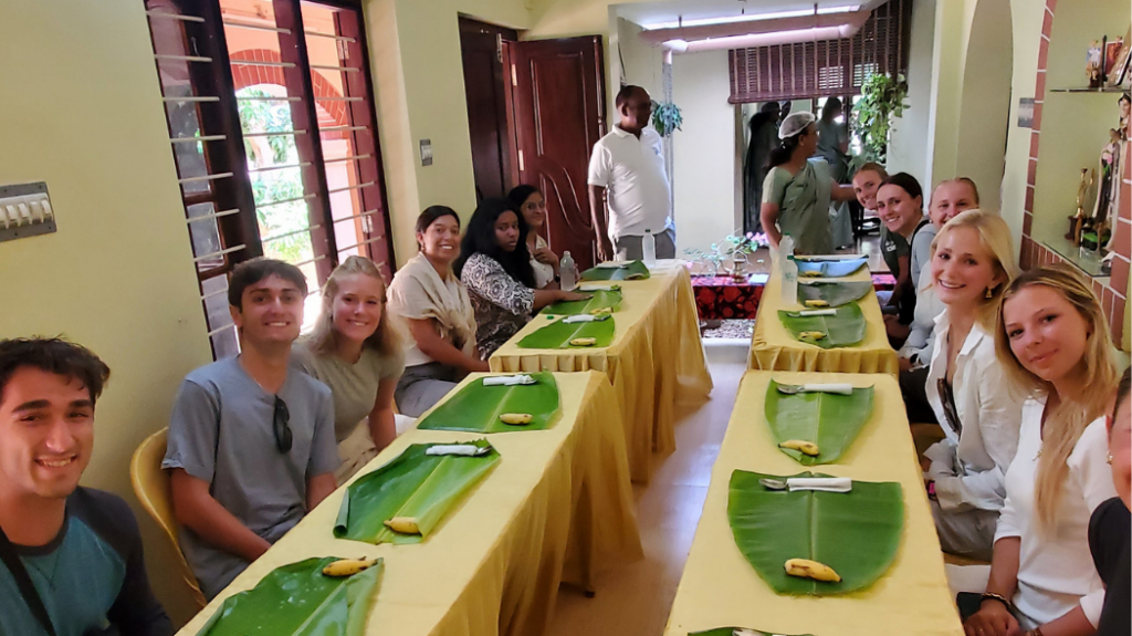 A group of people seated around several tables in a house in India, preparing for a meal.
