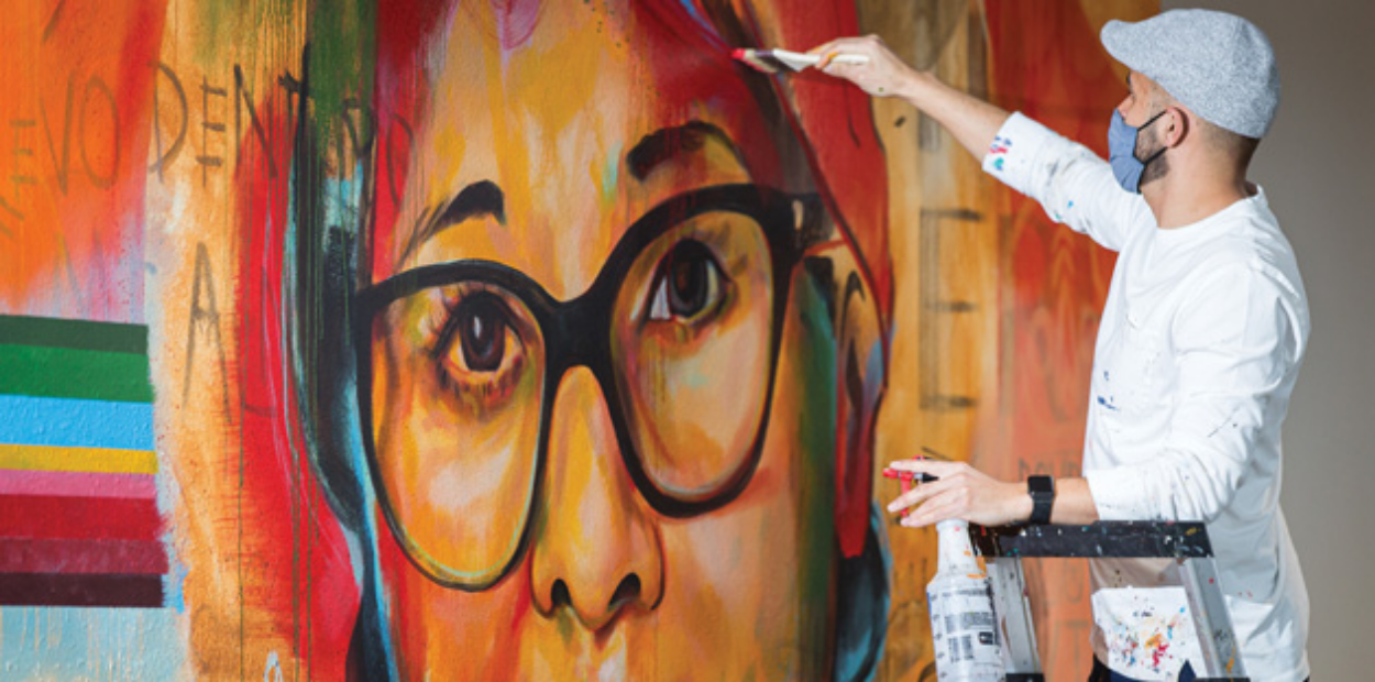 An artist wearing a face mask painting a colorful mural of a woman in glasses.
