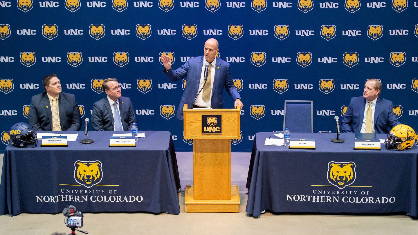 Sports administrator at podium talking to crowd with sitting administrators on either side.