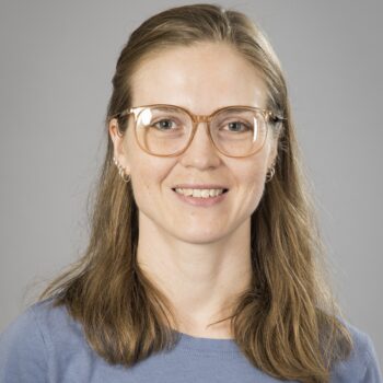 A person with long brown hair and glasses smiles warmly while wearing a blue t-shirt against a plain gray background.