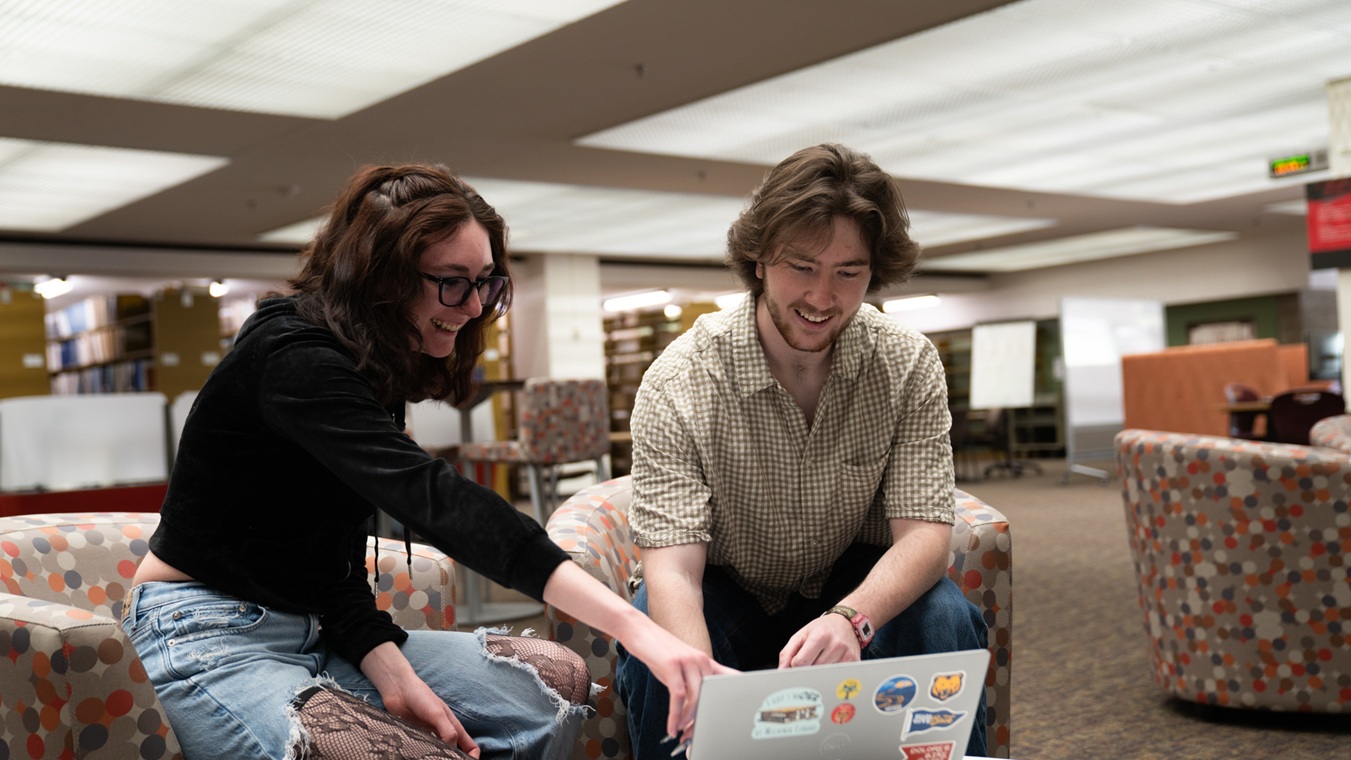 Two students seated in chairs looking at a laptop on a table in front of them. One student is pointing to the laptop screen.