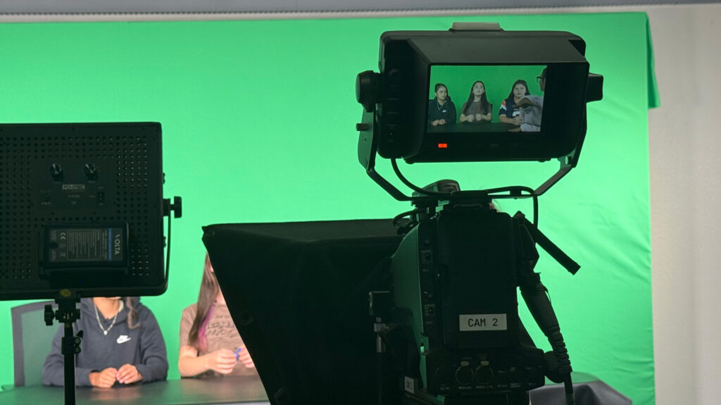 A camera screen showing three University Middle School students sitting at a desk in front of a green screen