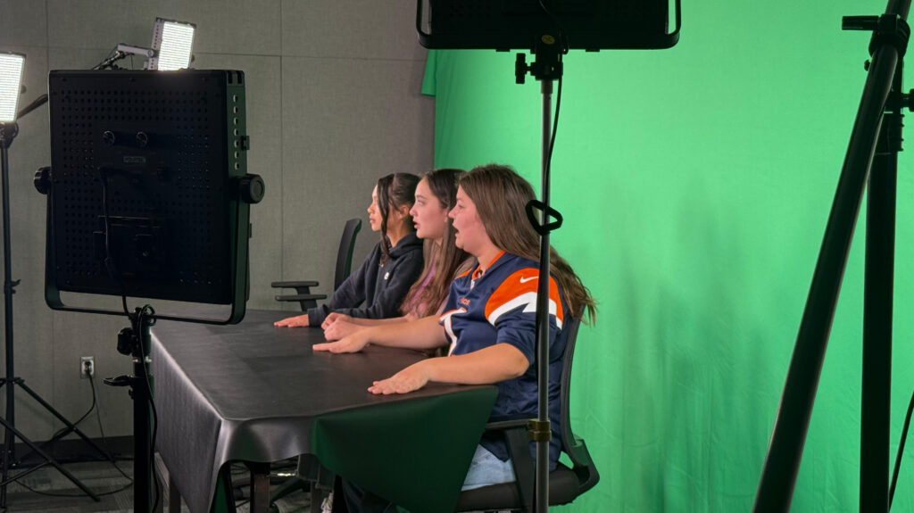 Three University Middle School students sitting at a desk in front of a green screen