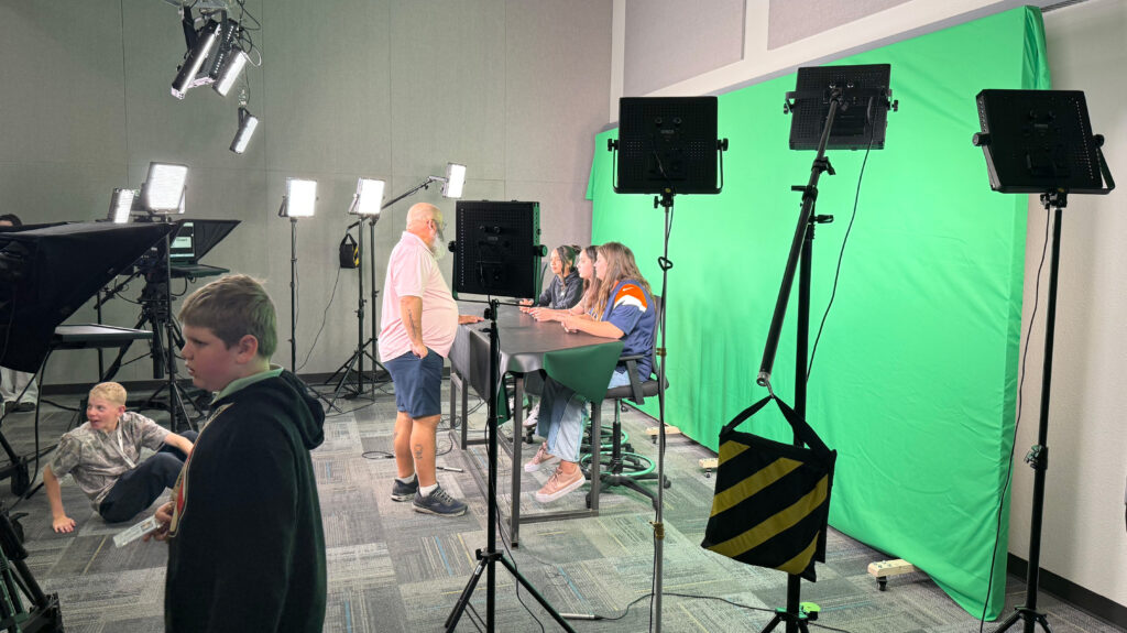 Three University Middle School students sitting at a desk in front of a green screen with cameras and lights around