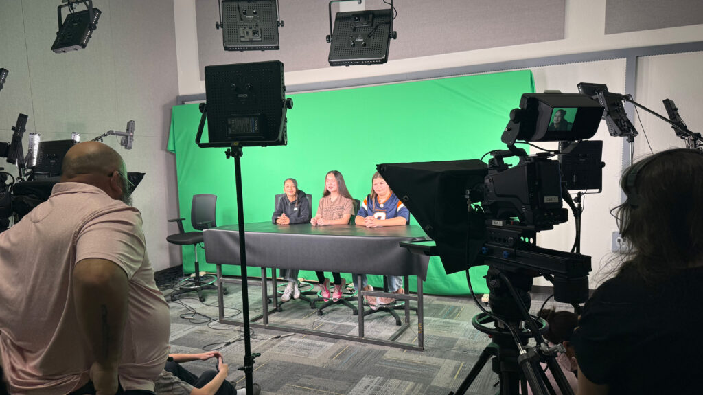 Three University Middle School students sitting at a desk in front a green screen