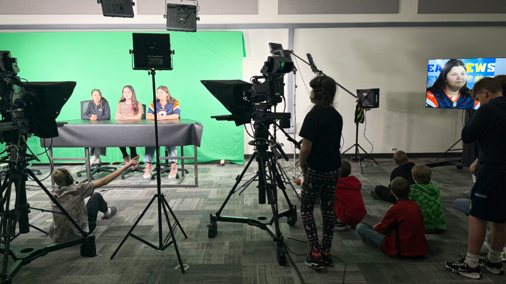 Three University Middle School students sitting at a desk with a green screen behind them, other students are sitting on the floor watching