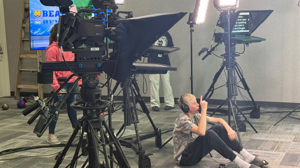 A middle school student sitting on the floor in front of cameras holing up a number 1