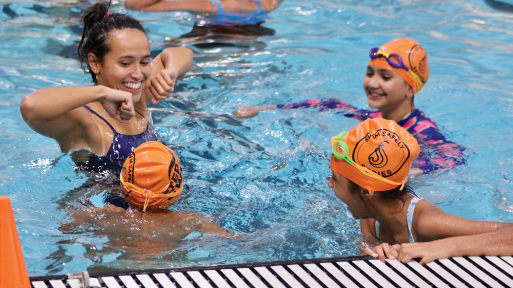 A UNC swim team member standing in the pool with three young swimming students.