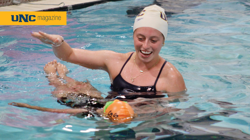 A UNC swim team member helping a young child to float on their back in a pool.