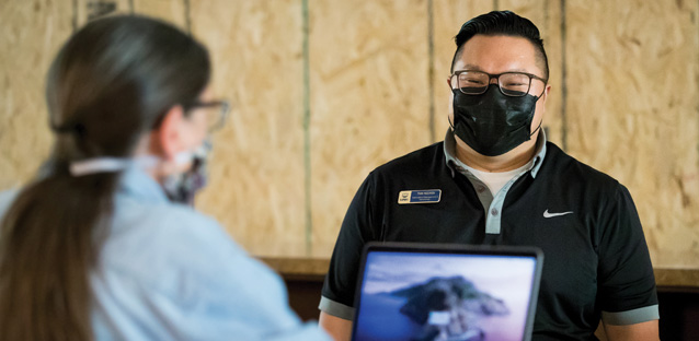 An IT worker wearing a black mask is talking to a woman behind a computer wearing a mask