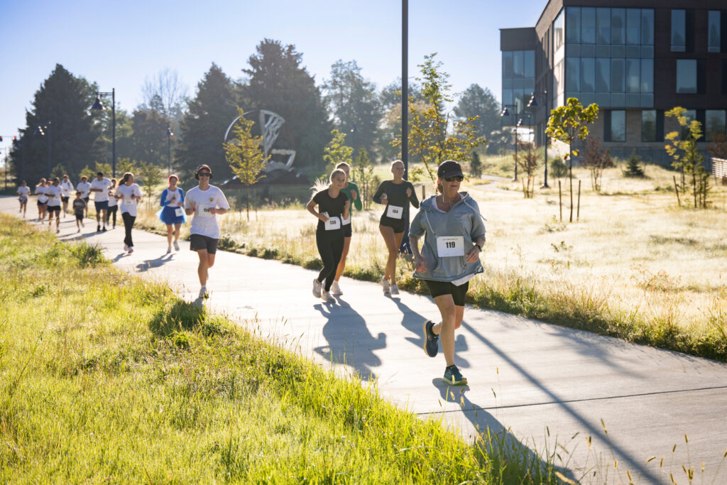 Runners with numbered bibs race along a sunlit paved path bordered by grass and trees, with modern buildings in the background.